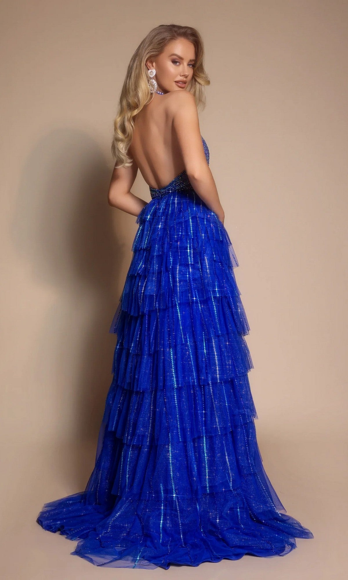 A woman stands against a beige background wearing the Long Formal Dress: Portia and Scarlett PS26117, a layered royal blue evening gown with sparkling beadwork, strapless and backless, paired with statement earrings.